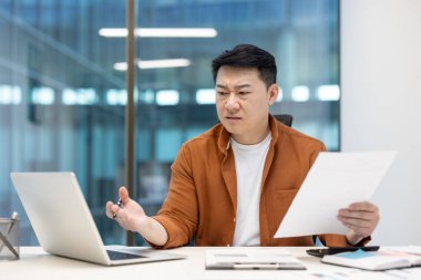 Ofiste dizüstü bilgisayarla dokümanları eleştirel bir ifadeyle inceleyen Asyalı adam, bir anlık profesyonel sorgulamayı yansıtıyor..