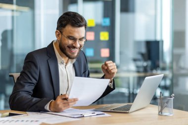 Olgun bir adam ofisinde oturmuş dizüstü bilgisayar kullanırken heyecanlı bir şekilde bir belgeyi inceliyor. Mesleki başarı ve motivasyonu temsil ederek, başarısı hakkında mutluluk ve kutlamaları ifade eder.