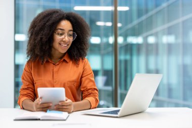 Profesyonel bir kadın dijital teknolojiyle ilgileniyor. Ofis ortamında tablet ve dizüstü bilgisayar da dahil. Modern çalışma alanında cam duvarlar var.