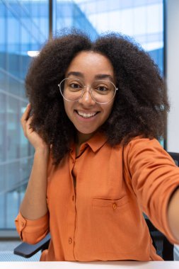 Gözlük takan ve iş yerinde selfie çekerken gülümseyen kendinden emin genç bir kadın. Görüntü doğal ışıklandırmayı, parlak bir atmosferi ve büyük cam pencerelerden görülebilen modern mimariyi gösteriyor.
