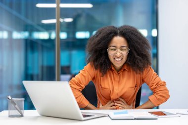 Ofiste çalışan bir kadın bariz bir şekilde mide ağrısı çekiyor. Gözlük ve turuncu gömlek takıyor, elleri karnını tutuyor, rahatsızlık veriyor..