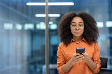 Ofis çalışanı gözlüklü ve turuncu tişörtlü profesyonel bir ortamda telefon kullanıyor. Cam ve ışıklarla modern arkaplan, teknoloji, iş ve odaklanma hissini iletiyor.