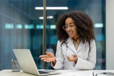 Ofisinde oturan ve bilgisayarından video konsültasyonu yapan bir kadın doktor. Profesyonel bir tavır sergiliyor ve hastalarla iletişim kurmak için sanal teknoloji kullanıyor..