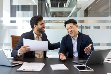 İki neşeli profesyonel modern bir ofis masasında oturmuş, canlı bir sohbet yapıyorlar. Çevre, belgeler, dizüstü bilgisayarlar ve çalışma faaliyetlerini gösteren olumlu bir işbirliği öneriyor