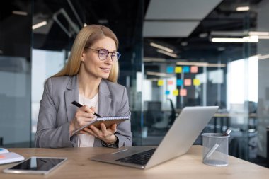 Modern ofis ortamındaki olgun kadın iş kadını, dizüstü bilgisayar üzerinde çalışırken not alıyor, profesyonelliği, kendini adamışlığı ve organizasyonu gösteriyor.