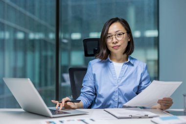 Modern ofis ortamında belgelerle ve dizüstü bilgisayarla çalışan Asyalı bir iş kadını. Profesyonel bir duruşu var. Odaklanma ve göreve olan bağlılığını açıkça gösteriyor..