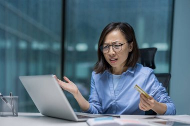 Modern bir ofisteki Asyalı iş kadını, elinde bir kredi kartı tutarken ve bilgisayarını kullanırken şaşkınlığını dile getiriyor. Finansal ya da teknik bir sorunla uğraşıyor gibi görünüyor..