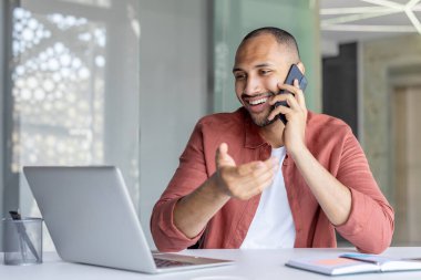 Neşeli bir profesyonel, uzaktan bilgisayarla çalışırken bir telefon görüşmesi yapar. Modern çalışma esnekliği ve işbirliği sergiler..