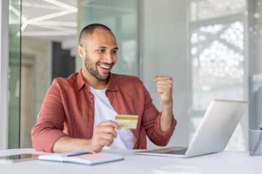 Neşeli bir adam, parlak bir ofis ortamında kredi kartı ve dizüstü bilgisayar kullanarak başarılı bir çevrimiçi işlemi kutluyor. Görüntü, dijital teknoloji ve memnuniyet kavramlarını iletir.