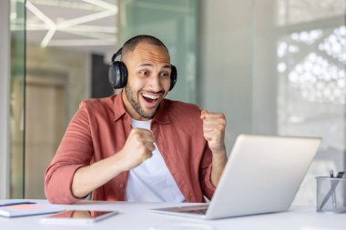 Kulaklık takan neşeli bir adam dizüstü bilgisayarı üzerinde çalışırken başarısını sevinçle kutlar. Modern ofis ortamında heyecan ve pozitif enerjiyi yakalamak.