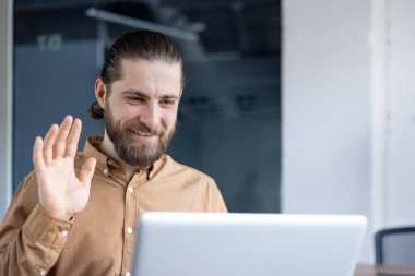 Sıradan gömlekli sakallı bir adam ekranda el sallayarak ve gülümseyerek bir video görüşmesine katılıyor. İletişim, uzak çalışma veya çevrimiçi profesyonel etkileşimler için ideal temsil.