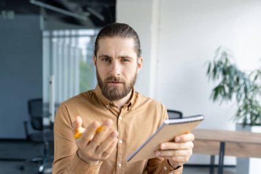 Focused professional man holding a notebook, looking judgmental, inquisitive, and serious, likely during a business interaction in an office environment clipart