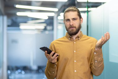 Modern bir ofis ortamında elinde akıllı bir telefonla duran ve kafa karışıklığını ifade eden bir adam. Mesleki kıyafet ve davranışlar, işyeriyle ilgili bir durum olduğunu gösteriyor..
