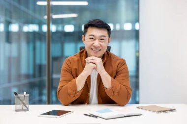 Yetişkin Asyalı iş adamı masa başında oturmuş, profesyonel bir ortamda güvenle gülümsüyor, ofis eşyalarıyla birlikte..