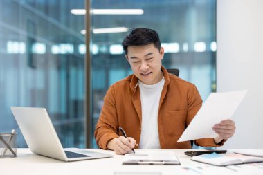 İşte çalışan Asyalı adam, gündüzleri ofiste notlar yazarken belgeyi gözden geçiriyor.