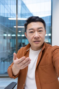 Yetişkin Asyalı iş adamı kameraya bakarak görüntülü konuşma yapıyor. İçerde, profesyonel bir ortamda, ciddi ve ilgi çekici bir iletişim kuruyor.