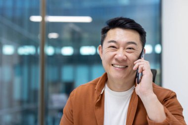 Asyalı bir adam telefonda gülümsüyor, parlak bir ofis ortamında oturuyor. İletişim kurarken kendine güvenme ve profesyonellik gösterme, mutluluk ve mutluluk gösterme