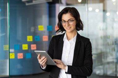 Modern ofis ortamında tablet kullanan kendine güvenen bir iş kadını görevlerle etkili bir şekilde meşgul oluyor. Arka planda parlak renkli yapışkan notlar takım çalışmasını ve yenilikçi fikirleri vurguluyor.