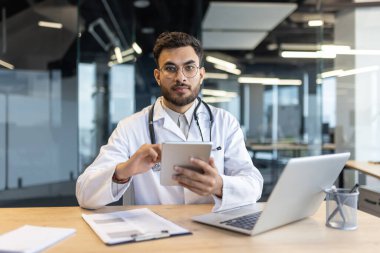 Beyaz önlüklü ve steteskoplu Latin Amerikalı doktor profesyonel bir ortamda tablet ve dizüstü bilgisayarla çalışarak, sağlık ve teletıp hizmetlerinde modern teknoloji kullanımını göstermektedir..