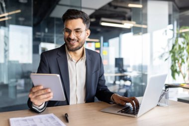 Latin Amerikalı işadamı modern bir çalışma alanında tablet ve dizüstü bilgisayar kullanarak mesleki bağlılık ve verimliliği gösteren görevler üzerinde çalışıyor..