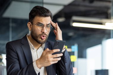 Takım elbiseli Latin Amerikalı bir işadamı, ofisteki akıllı telefonuna bakarken şok geçiriyor..