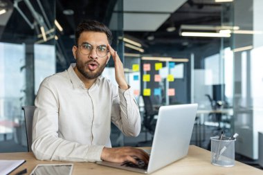 Latin Amerikalı bir işadamı, modern ofis ortamında beklenmedik haberler ya da keşifler sunan bir dizüstü bilgisayar kullanarak şaşkınlığını dile getiriyor. Çevre verimliliği ve mesleki işi yansıtıyor