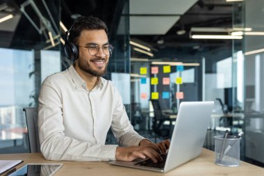 Latin American man using a laptop while wearing headphones in a modern office environment. Captures a professional setting, teamwork planning in the background, and focus on productive work.