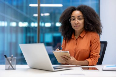 İş yerindeki ciddi bir iş kadınının portresi, iş kadını kameraya bakıyor, dizüstü bilgisayarla çalışıyor, elinde bir defter tutuyor, online bir toplantıdan veri kaydediyor.