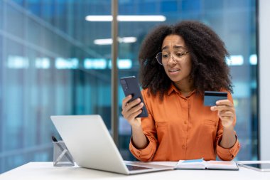 İş yerindeki üzgün ve hayal kırıklığına uğramış kadın, elinde telefon ve banka kartı ile çevrimiçi para transferi, dolandırıcılık eylemleri ve hesabı engellemeye çalışıyor..