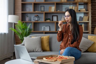 Asyalı kadın evde dizüstü bilgisayar izlerken pizza yemekten hoşlanır. Rahat ve rahat atmosfer, rahatlık, teknoloji ve rahatlığı ön plana çıkarıyor. Modern yaşam tarzının mükemmel tasviri.