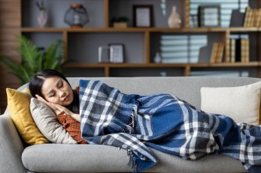 Asyalı kadın kanepede huzur içinde dinleniyor, rahat bir battaniyeye sarılıp, ev ortamında rahatlık ve rahatlık hissi uyandırıyor. Huzur ve huzur dolu yaşamın mükemmel tasviri..