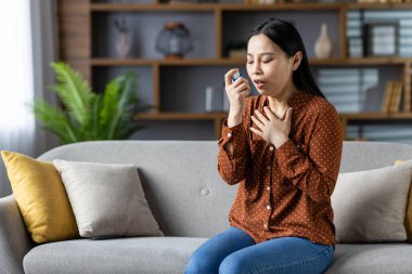 Astım spreyi kullanan Asyalı bir kadın, kanepede oturuyor ve nefes alma sorunu yaşıyor. Puantiyeli bluz giymek, evde solunum sağlığını yönetmek. Odaklanmış, sakinleştirici atmosfer.