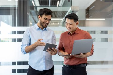 Gündelik giysiler içinde iki programcı yeni bir yazılım projesini tartışıyorlar. Pencerenin yanında dizüstü bilgisayarla duran, gülümseyen ve konuşan adamlar..
