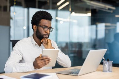 Ofisin içinde dizüstü bilgisayar ve tablet bilgisayarla çalışan erkek düşünen işadamı, iş gömlekli adam çevrimiçi mali raporu izliyor, monitör ekranından okuyor..