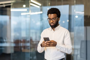 Ofisin içinde elinde telefonla duran ciddi odaklı bir adam, iş adamı düşünceli bir şekilde mesaj yazıyor, iş kıyafetleri giymiş bir çalışan iş yerinde uygulama kullanıyor..