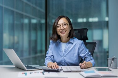 Asyalı genç muhasebecinin portresi. Evrakların arkasındaki iş kadını gülümsüyor ve kameraya bakıyor, iş yerindeki sözleşmeler ve faturalarla çalışıyor, vergi formlarını dolduruyor..