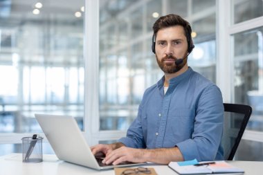 Ofis içinde kulaklık takmış kendine güvenen bir adamın portresi. Kameraya bakan destek çalışanı video görüşmesi için dizüstü bilgisayar kullanıyor. İnternet üzerinden müşteri hizmetleri danışmanlığı.