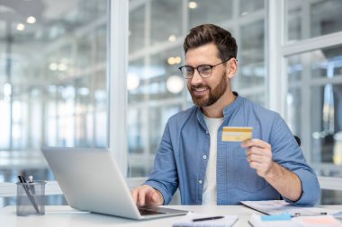 Bilgisayarlı ve kredi kartlı bir ofis çalışanı ofis içi masasında oturuyor. Bankacılık uygulaması kullanarak internete para aktarıyor. İş adamı gülümsüyor ve online rezervasyondan memnun.