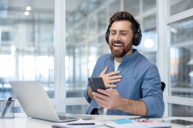 Kulaklıklı bir adam akıllı telefondan komik ve eğlenceli videolar izliyor, ofis çalışanı ofiste dizüstü bilgisayarla otururken gülüyor..