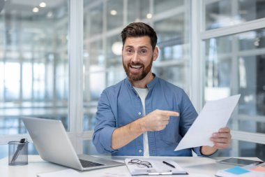 Başarılı bir finansörün portresi, elinde mali raporla kameraya bakan bir adam. Ofisin içindeki belgelerle çalışan memnun iş adamı..