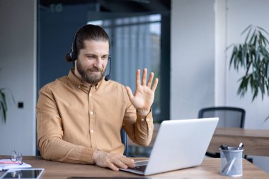 Kulaklık takan bir adam dizüstü bilgisayarla etkileşime geçiyor. Profesyonel bir ofis ortamında otururken el kol hareketi yapıyor..