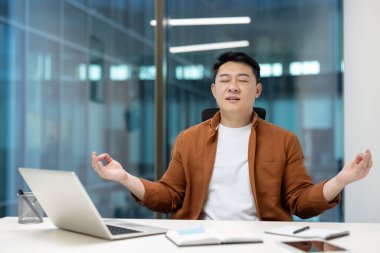 Olgun Asyalı adam ofiste Lotus pozisyonunda meditasyon yapıyor. İş yerinde dizüstü bilgisayarla çalışan iş adamı, nefes egzersizleri yapıyor..