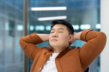 Asyalı adam dinleniyor ve yakından dinleniyor. Ofiste çalışan bir iş adamı gözleri kapalı ve elleri başının arkasında..