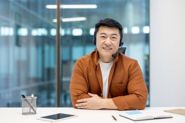 Webcam görüntüsü, kulaklıklı başarılı adam ciddi ve kameraya odaklı, Asyalı destek çalışanı ofisin kalbinde..