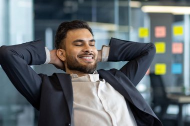 İyi yapılmış bir adam elleri başının arkasında dinlenir, hayaller kurar ve başarısının gelecek sonuçlarını düşünür. Ofisteki işyerinde çalışan bir işadamı iş elbisesiyle yakın plan çalışıyor.