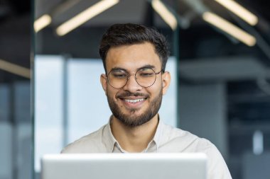 İşyerinde dizüstü bilgisayarla gülümseyen bir adamın yakın çekimi. İş adamı, iş gömleği ve gözlükleriyle klavyede daktilo kullanıyor, iş yerinde ofiste çalışıyor..