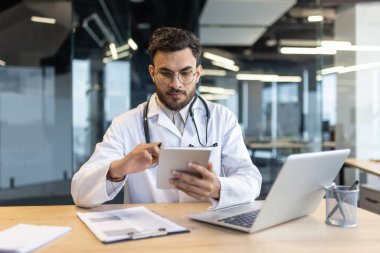 Masada dizüstü bilgisayarla oturan odaklanmış bir doktor. Beyaz önlüklü bir adam işyerindeki hastalara danışmanlık yapıyor..