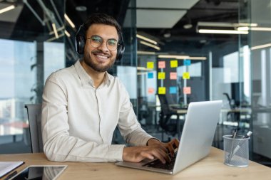 İş yerindeki bir ofiste dizüstü bilgisayarlı bir adamın portresi, kulaklıklı bir iş adamı gülümseyip kameraya bakıyor. Çevrimiçi çalışan ofis çalışanı.