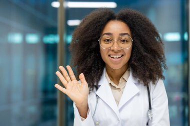 Çevrimiçi video danışmanlığı, genç başarılı bayan doktor kameraya bakar ve elini sallar, hastalarla uzaktan iletişim kurar. Webcam manzaralı, beyaz önlüklü kadın, steteskoplu..