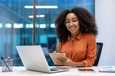 İşteki bir kadının portresi. Başarılı bir ofis çalışanı, gülümseyen ve kameraya bakan, notlar için bir not defteri tutan. Video görüşmesi ve çevrimiçi video konferansı için dizüstü bilgisayarı olan iş kadını.